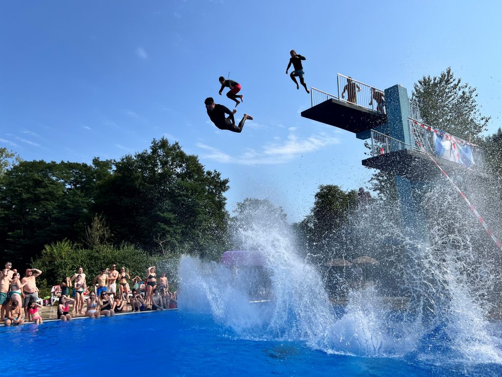 Mehrere Personen springen vom Sprungturm in Schwimmbecken. Um das Becken herum sitzen viele Personen